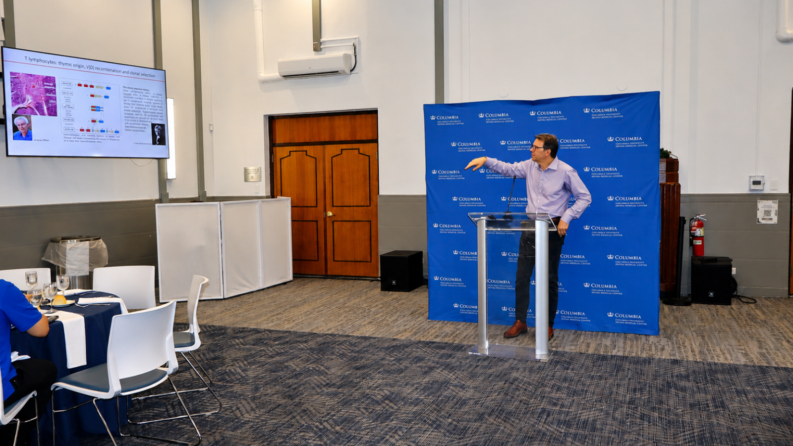 A presenter stands at a podium and points toward a presentation screen during an academic lecture at Columbia University Irving Medical Center.