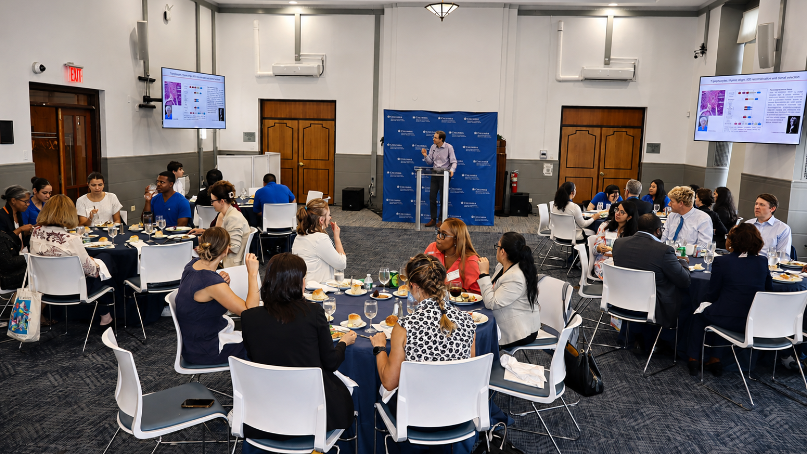 A presenter speaks at a podium during a CUIMC event as attendees sit at round tables, with research presentation slides displayed on screens throughout the room.