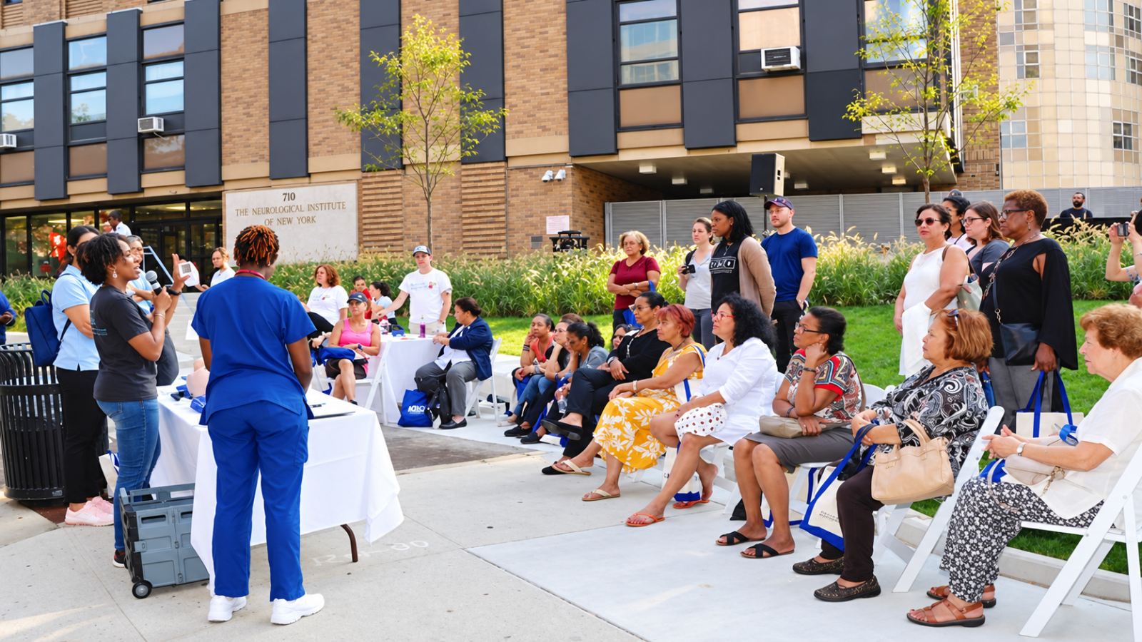 Community members and CUIMC staff gather outdoors for a health education event, with attendees seated and listening to presenters in front of the Neurological Institute of New York.