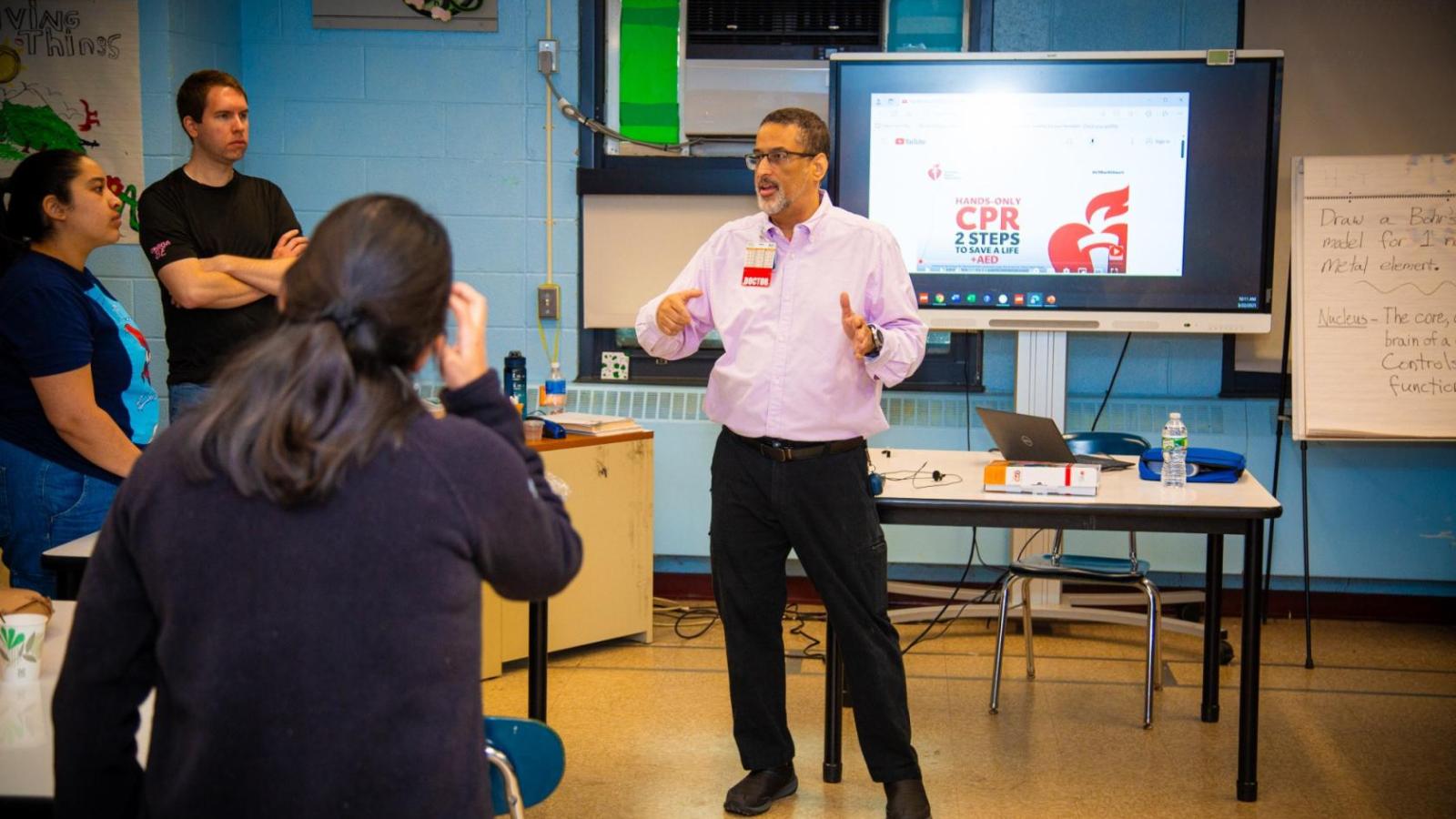 A physician speaks to participants during a hands-only CPR training session, with a CPR and AED presentation displayed behind him.