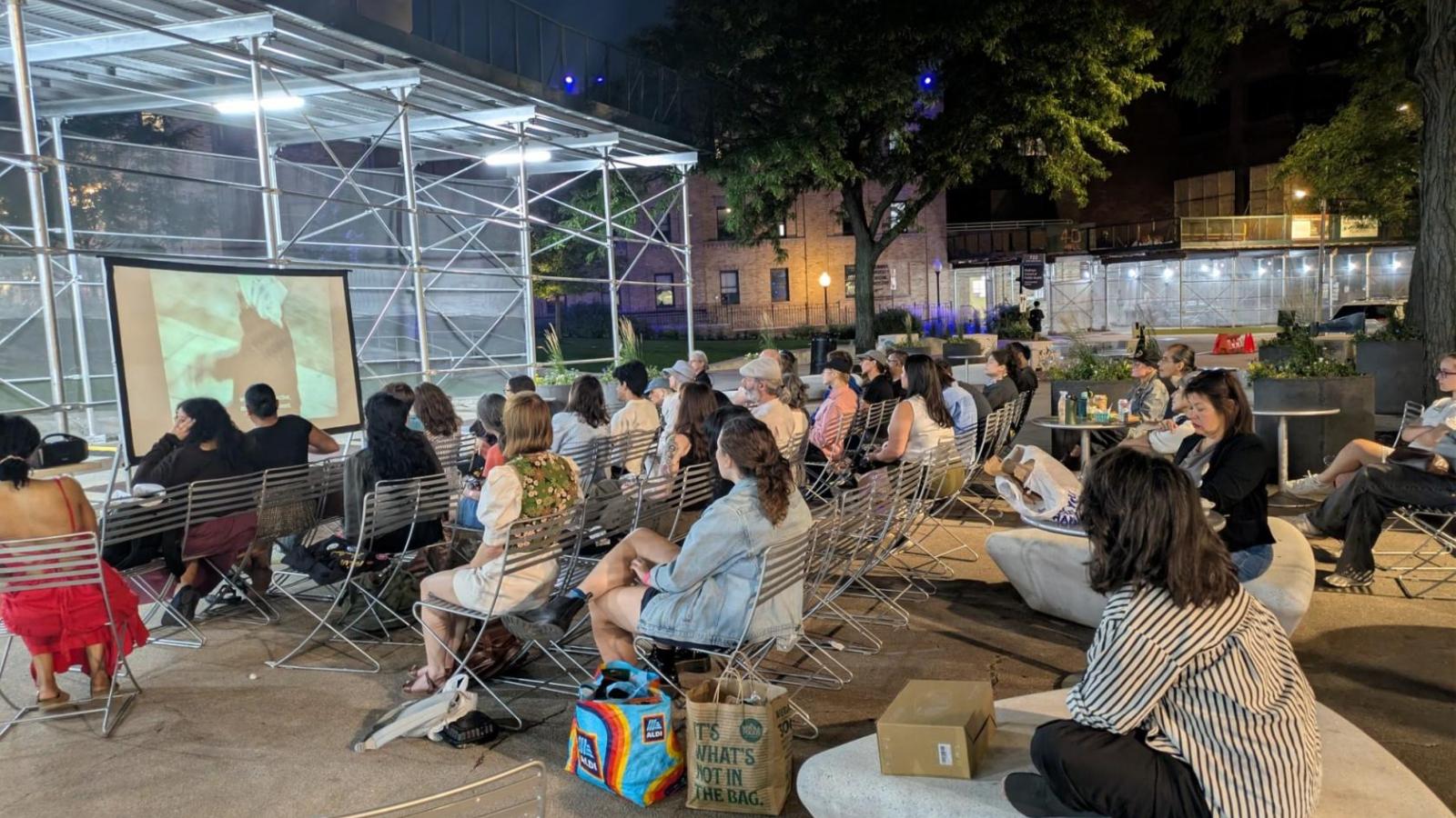 Audience members sit outdoors at Haven Plaza at night, watching a film projected on a large screen.