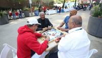 Four men sitting outdoors playing dominoes and laughing together at a community event.