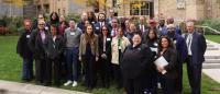 A large, diverse group of adults stands together outdoors on a grassy plaza, posing for a group photo. They are dressed in business and business-casual attire and wear name tags. Behind them are brick and concrete institutional buildings, steps, and lands
